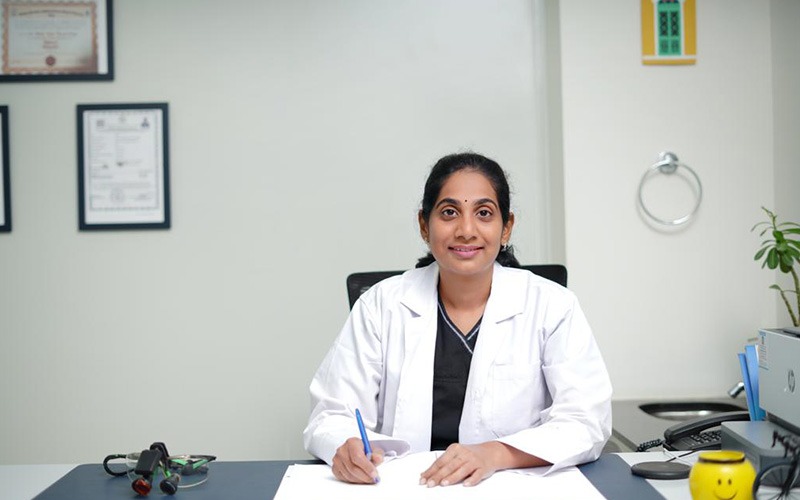 Dr. Sravanthi seated at her desk at the Tooth Matters practice in Kondapur, in a white coat, with framed qualifications on the wall behind her
